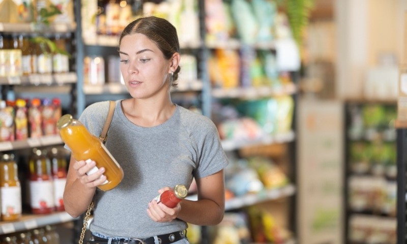 Zainteresovana mlada devojka u sivoj majici i farmerkama drži i ispituje flaše organskog soka
